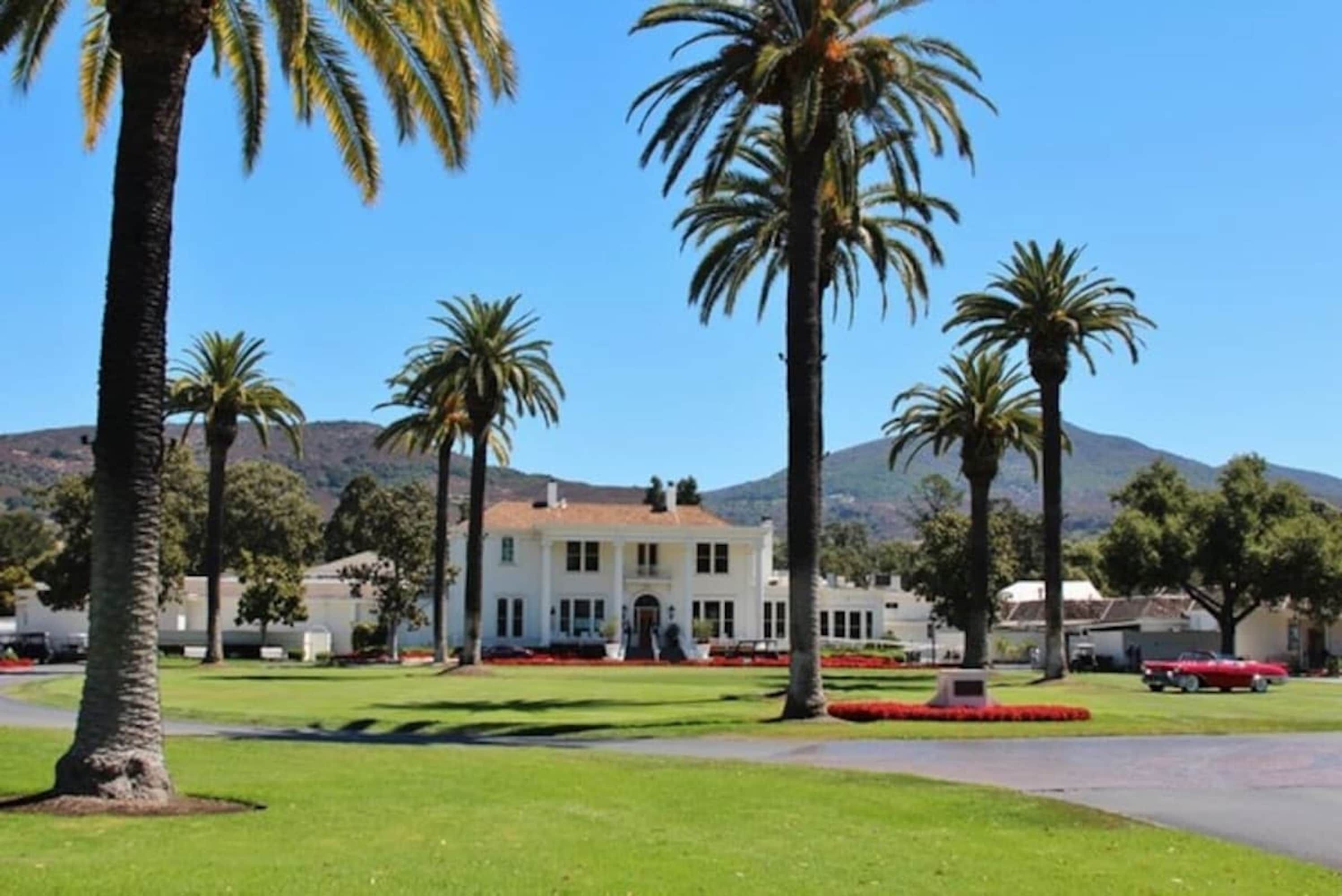 A large white building with a symmetrical facade, surrounded by well-maintained lawns and palm trees, set against a backdrop of hills.
