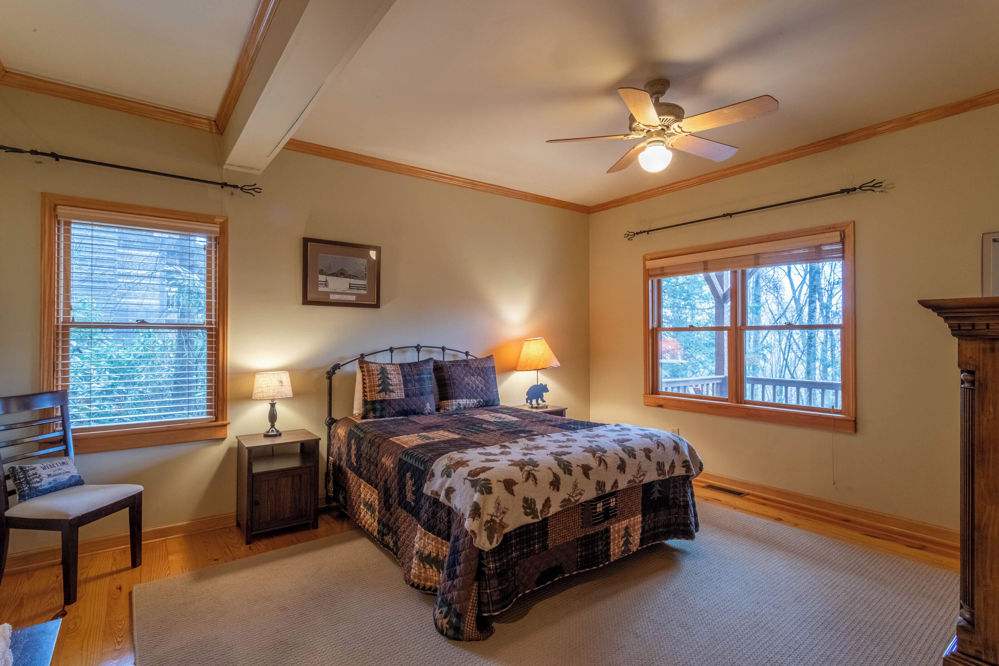 A bedroom with a bed, a chair, a nightstand, a lamp, a ceiling fan, and two windows.
