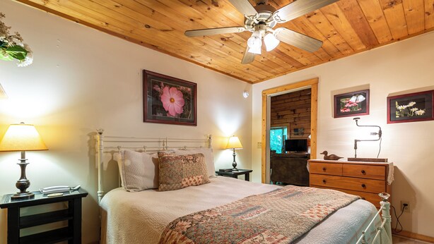 A bedroom with a wooden ceiling, a bed with a patterned blanket, two bedside lamps, a wooden dresser, and framed pictures on the wall. - Trillium (Banner Elk)