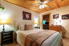 Ein Schlafzimmer mit Holzboden, einem Bett mit gemustertem Überwurf, zwei Nachttischlampen, einer Holzkommode und gerahmten Bildern an der Wand.