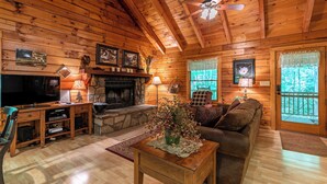 A cozy cabin interior with a stone fireplace, wooden walls, a ceiling fan, and a television. - Trillium (Banner Elk)