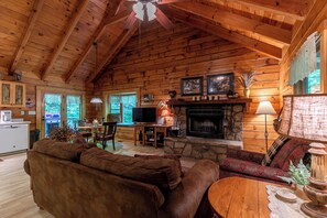 A cozy cabin interior with a stone fireplace, wooden ceiling beams, a sofa, and a television. - Trillium (Banner Elk)