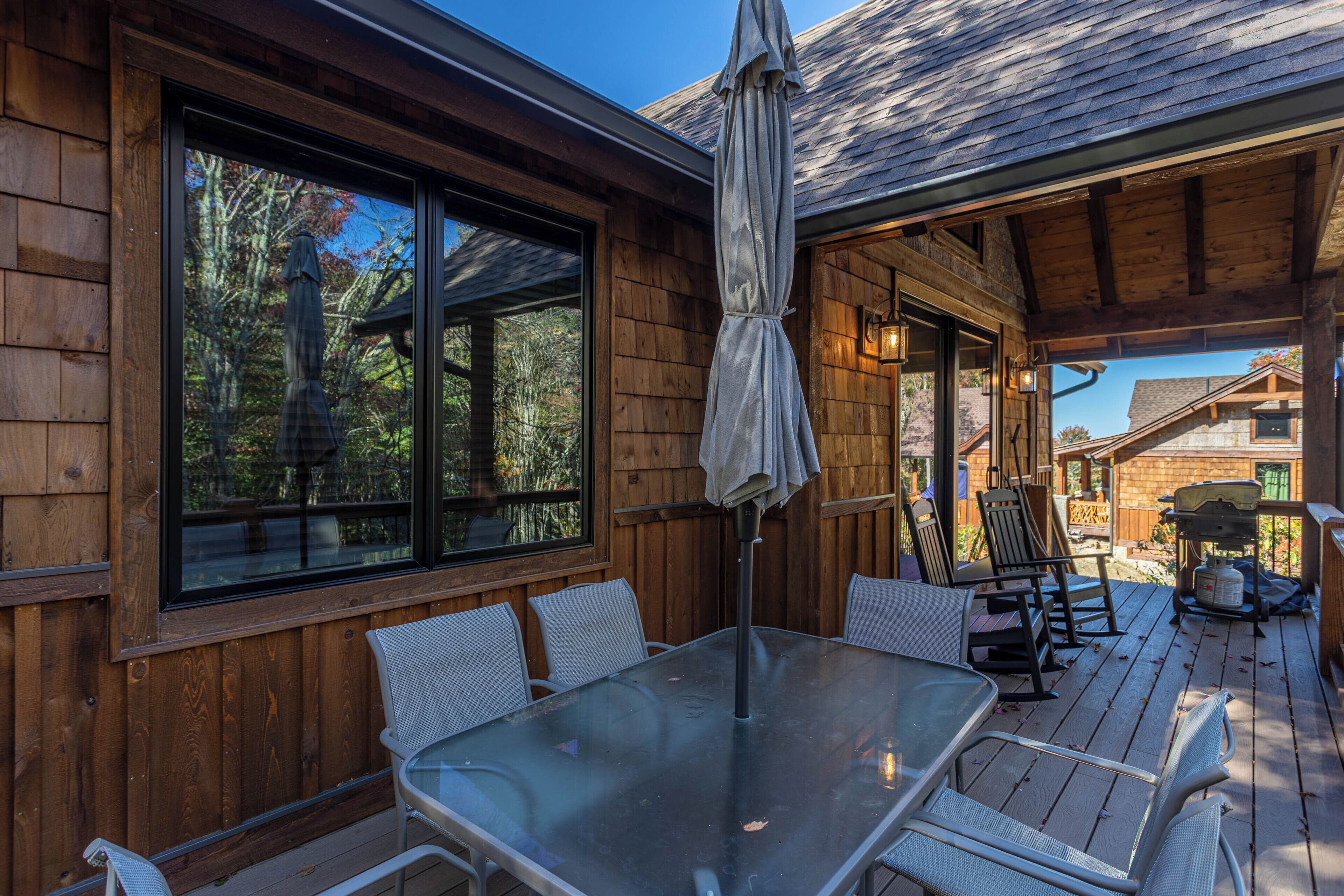 A wooden deck with a glass table and chairs, a covered seating area, and a view of the surrounding forest.