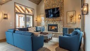 A cozy living room with a stone fireplace, a flat-screen TV mounted above it, and a large window providing natural light. - Birds Nest Retreat at Eagles Nest (Banner Elk)