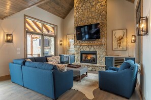 A cozy living room with a stone fireplace, a flat-screen TV mounted above it, and a large window providing natural light. - Birds Nest Retreat at Eagles Nest (Banner Elk)