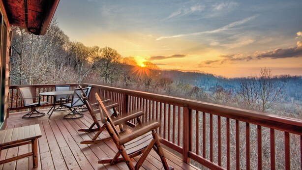 A wooden deck with outdoor furniture, overlooking a forested landscape with a sunset view. - Sugars Sunset Vista (Banner Elk)