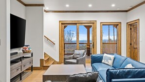 A modern living room with a TV, sofa, chairs, and a staircase leading to a balcony with a view. - The Dire Wolf at Eagles Nest (Elk Park)
