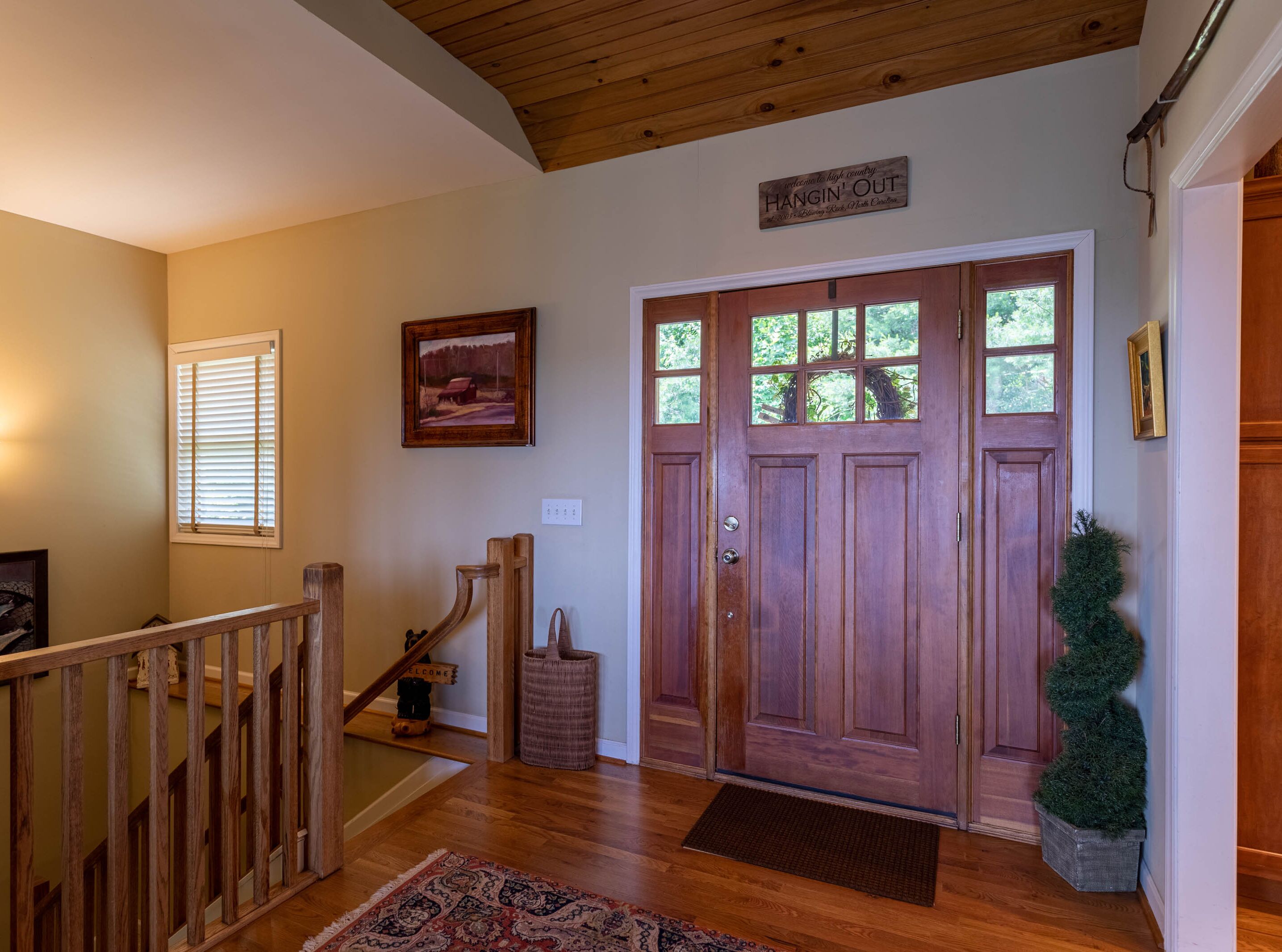 A wooden staircase leading to a door with a welcome sign, a framed landscape painting, and a small Christmas tree.
