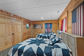A room with wooden walls, a large bed with a quilt, a door with an American flag, and a window with a curtain. - Falling Waters (Banner Elk)