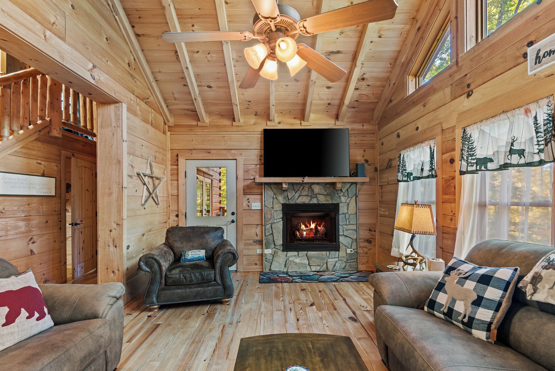 A cozy cabin interior with a stone fireplace, wooden walls, a ceiling fan, and a television mounted above the fireplace.