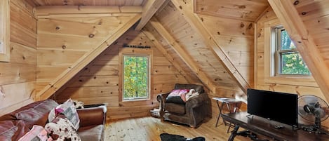 A cozy attic room with wooden walls and a sloped ceiling. There is a leather couch, a television, and a fan.
