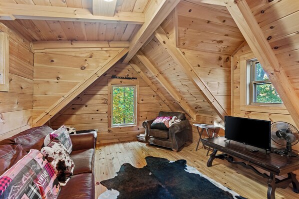 A cozy attic room with wooden walls and a sloped ceiling. There is a leather couch, a television, and a fan. - Falling Waters (Banner Elk)