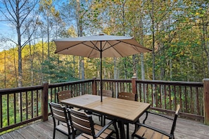 A wooden deck with a table and chairs, surrounded by a wooden railing, overlooking a forested area. - Falling Waters (Banner Elk)