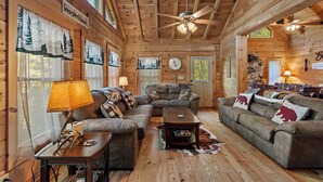 A cozy cabin interior with a large wooden ceiling, a ceiling fan, a fireplace, and a dining area. - Falling Waters (Banner Elk)