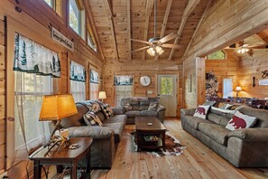 A cozy cabin interior with a large wooden ceiling, a ceiling fan, a fireplace, and a dining area. - Falling Waters (Banner Elk)