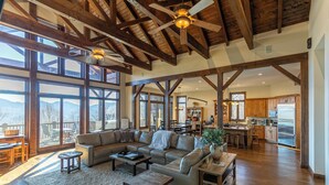 A spacious living room with a large sectional sofa, wooden beams, and large windows offering mountain views. - Banner Crest - Mountain Luxury! Views, hot Tub, Private Pool and Tennis Courts, Game Room (Banner Elk)