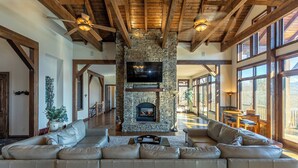A spacious living room with a stone fireplace, leather sofas, and large windows offering views of the outdoors. - Banner Crest - Mountain Luxury! Views, hot Tub, Private Pool and Tennis Courts, Game Room (Banner Elk)