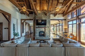 A spacious living room with a stone fireplace, leather sofas, and large windows offering views of the outdoors. - Banner Crest - Mountain Luxury! Views, hot Tub, Private Pool and Tennis Courts, Game Room (Banner Elk)
