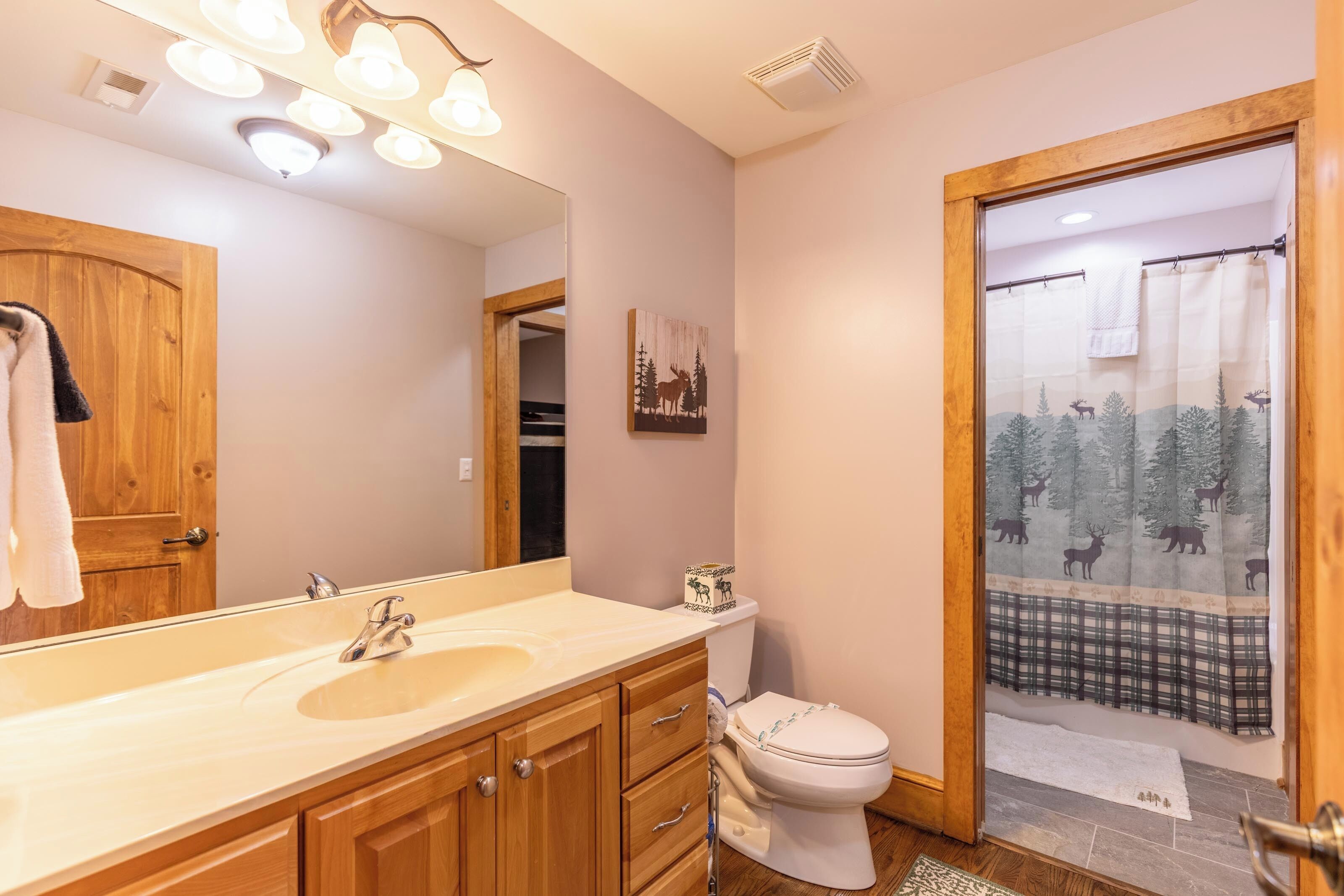 A bathroom with a wooden vanity, a large mirror, a toilet, and a shower curtain with a forest design.