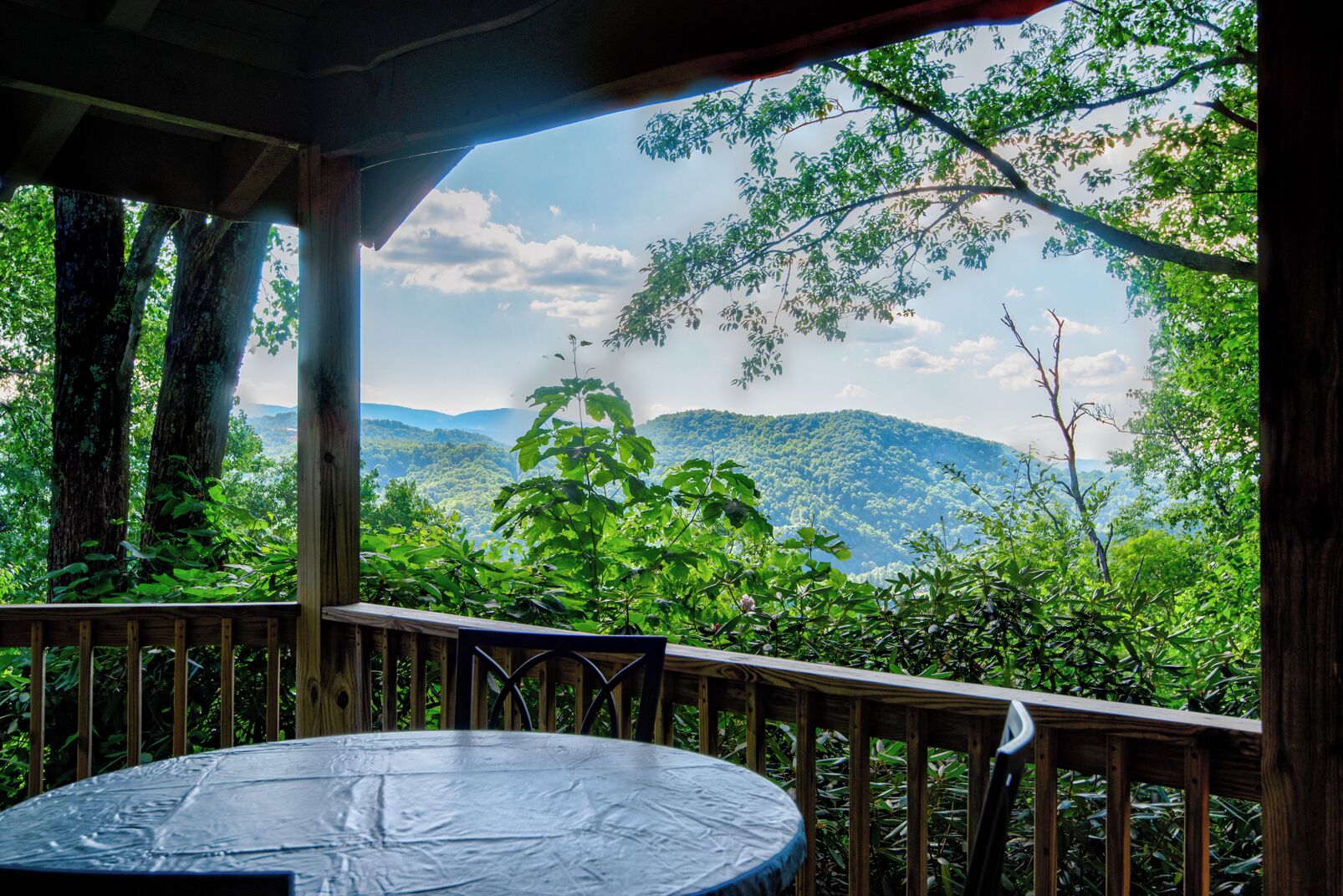 A wooden deck with a table and chairs, overlooking a lush forest and distant mountains.