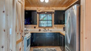 Una cocina compacta con paredes de madera, estufa integrada, armarios negros y un refrigerador de acero inoxidable, además de una ventana con persianas.