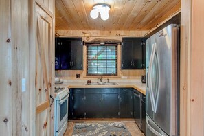 A compact kitchen with wooden walls, a built-in stove, black cabinets, a stainless steel refrigerator, and a window with blinds. - Crystal View (Boone)