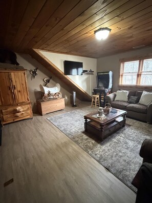 A loft-style living room with a sloped ceiling, wooden beams, a television mounted on the wall, a gaming console, a sofa, and a coffee table. - Country Life (Vilas)