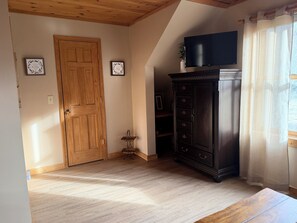 Una habitación con puerta de madera, un armario de madera oscura, un televisor de pantalla plana y una ventana con cortinas translúcidas.
