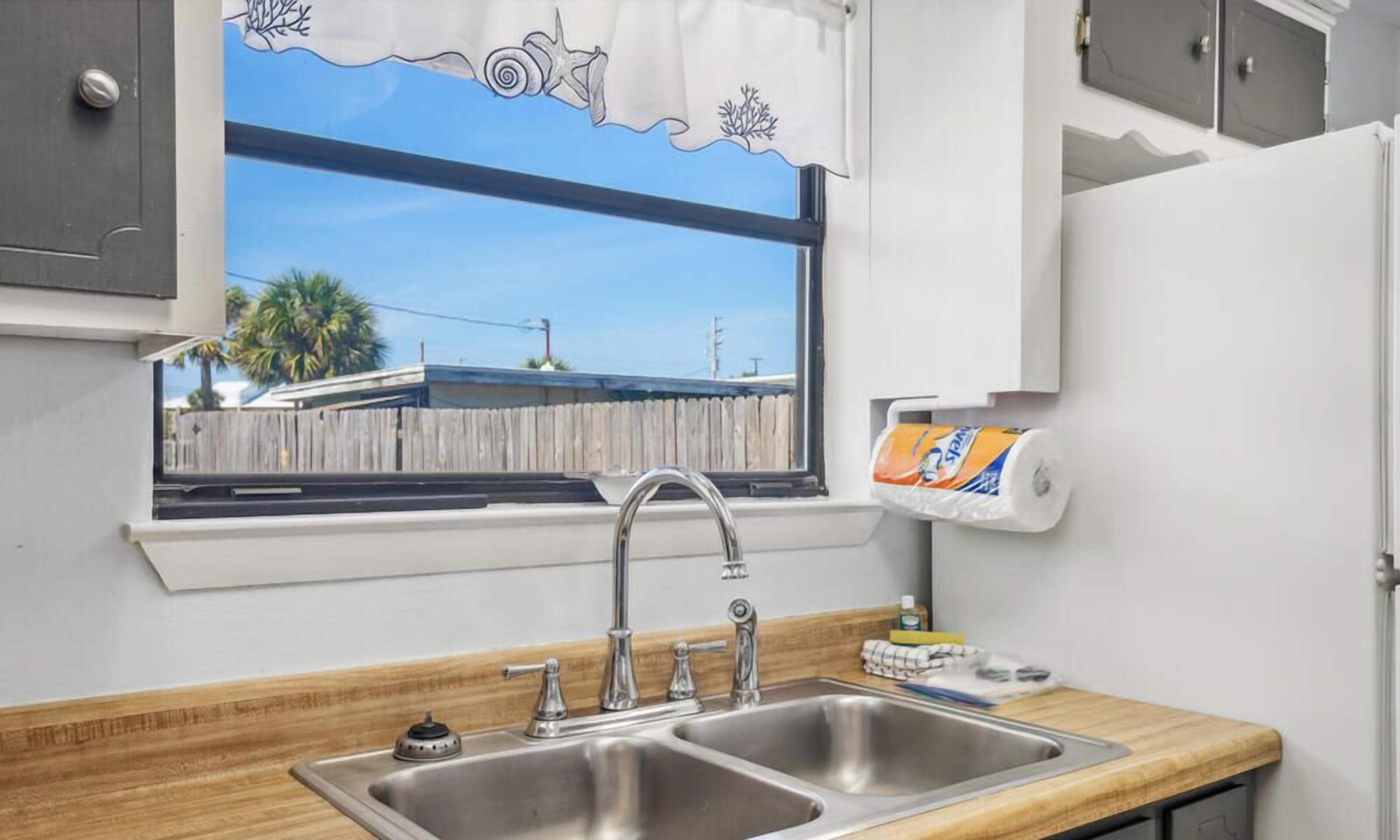 A kitchen with a stainless steel sink, a window with a view of a wooden fence and palm trees, and a paper towel dispenser.