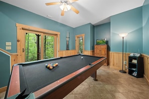 Une table de billard avec des billes, un ventilateur de plafond et des fenêtres donnant sur la forêt.