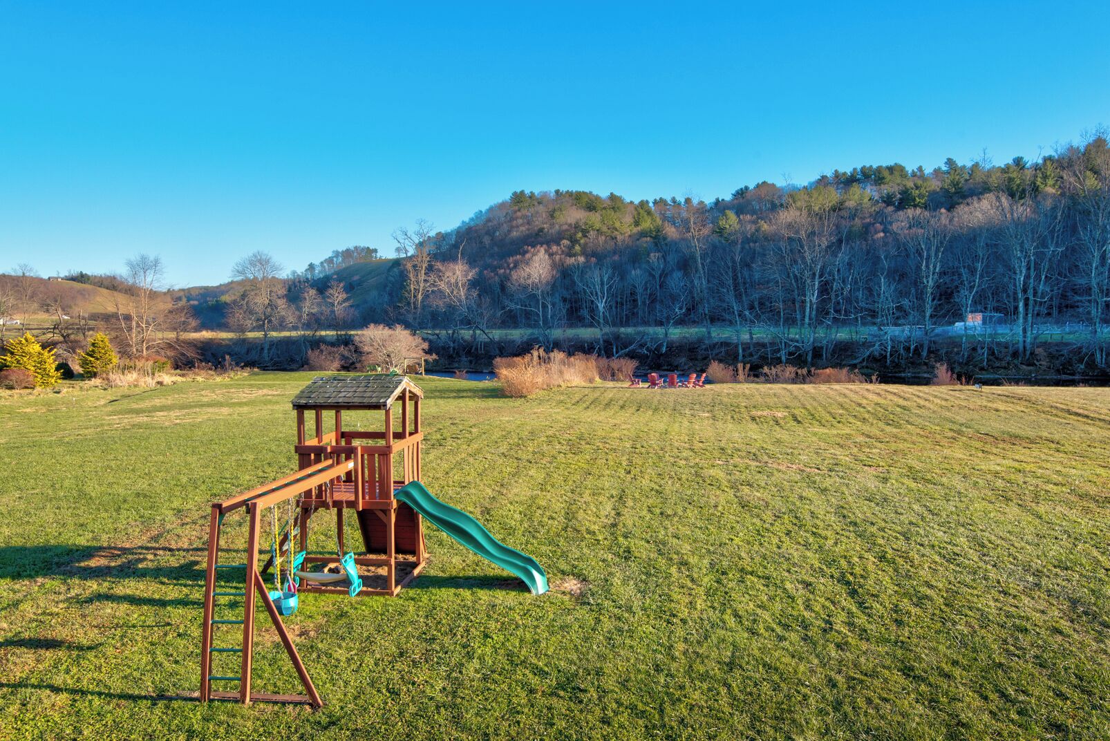 A playground with a slide and climbing structure in a grassy field with a forested hill in the background.
