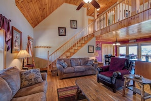 A spacious living area with a wooden staircase, a dining area with a table and chairs, and a view of the outside through large windows.