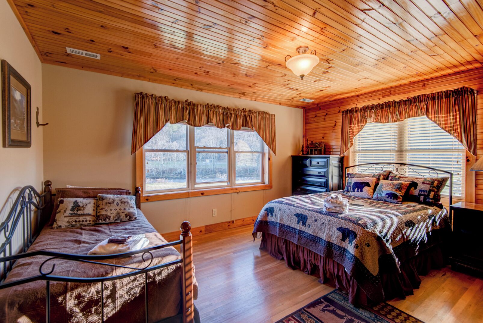 A cozy bedroom with wooden ceilings, a large bed with a patterned blanket, a smaller bed, and a dresser.