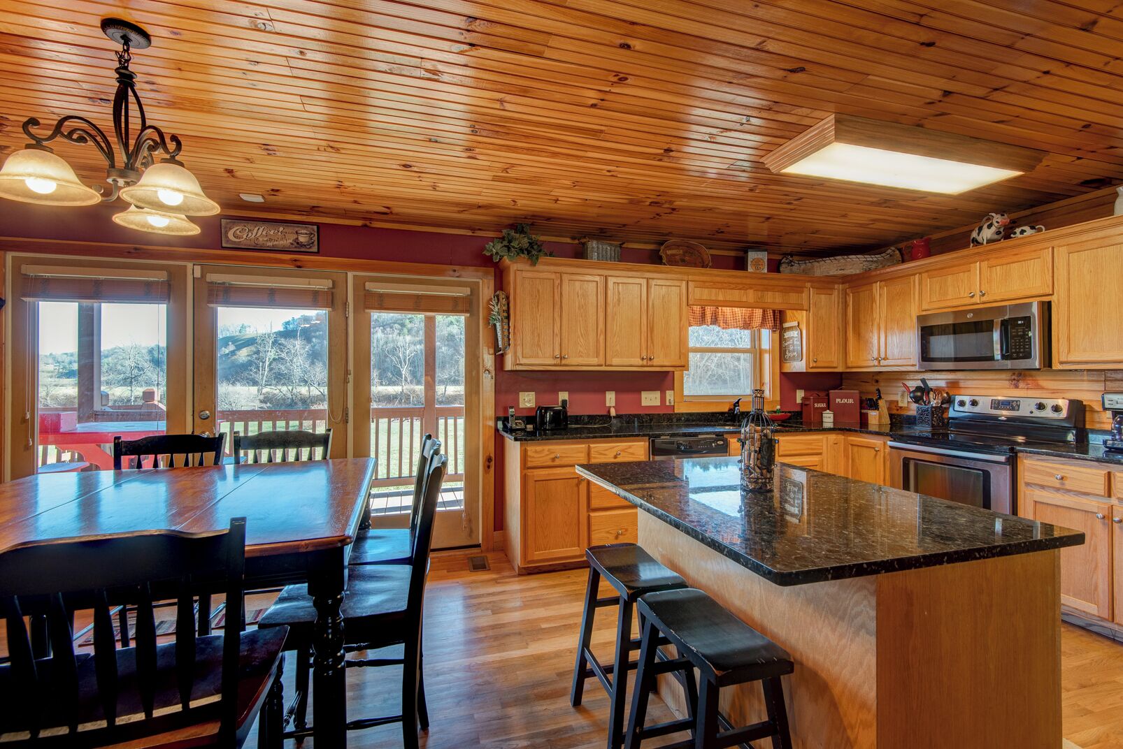 Una cocina amplia con armarios de madera, encimera de granito y una isla grande. Hay un comedor con mesa y sillas, y vistas al exterior a través de ventanas grandes.
