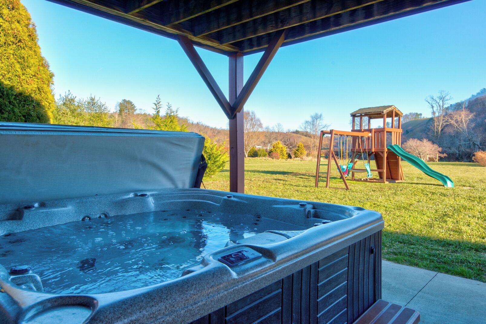 A hot tub under a wooden pergola with a playground in the background.