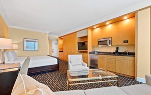 Una habitación de hotel con cama, sofá, una mesa de centro de vidrio, televisor y una kitchenette con microondas y cafetera.
