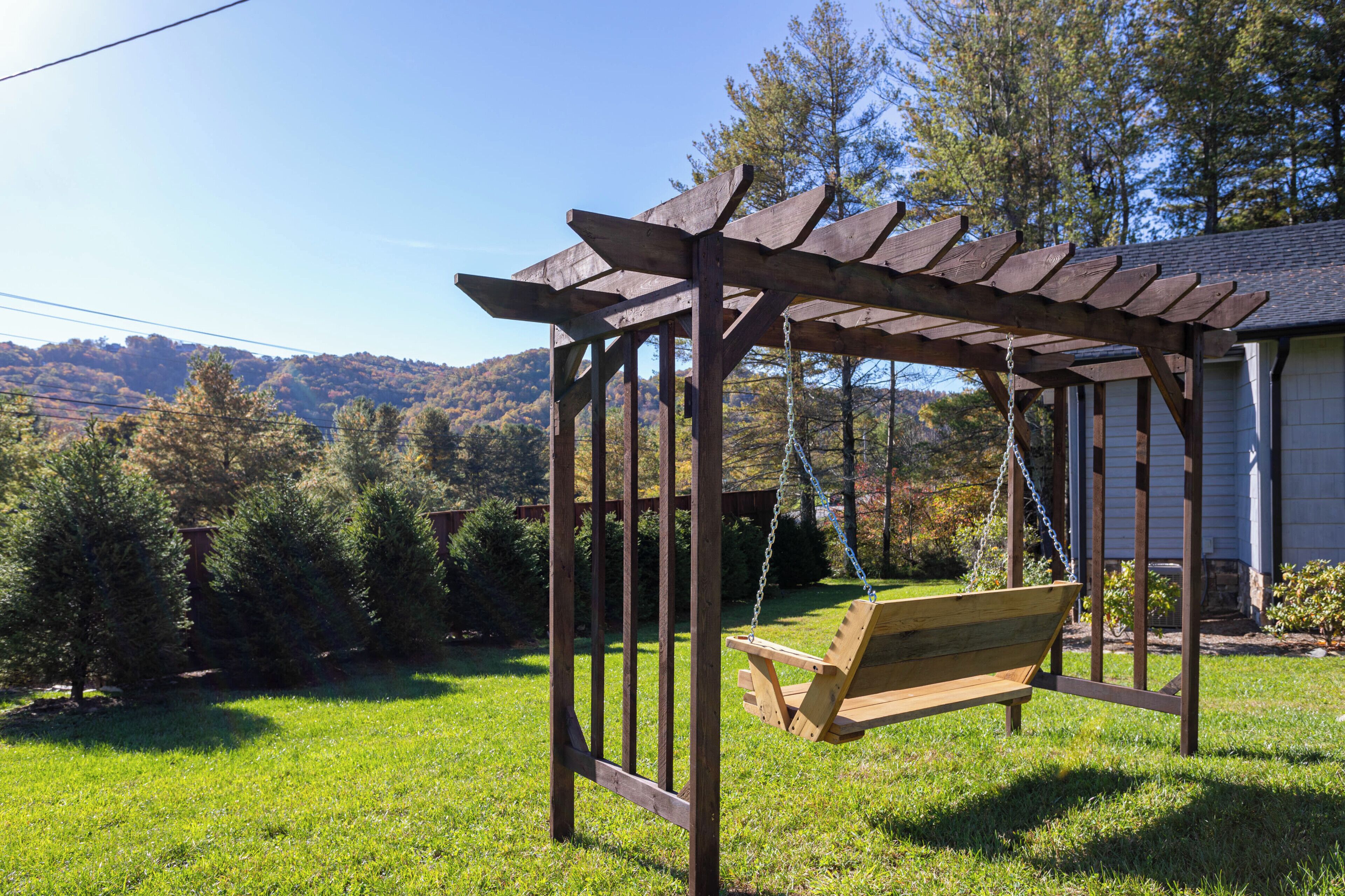 A wooden swing set with a bench, surrounded by a grassy yard and trees, with a house in the background.