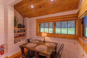 A dining area with a wooden table, chairs, a window with a view of a grassy field, and a shelf with decorative items. - Banner Sky (Banner Elk)