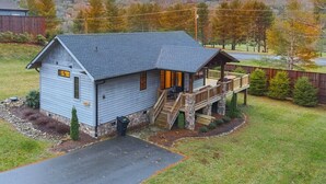 Ein Bungalow mit Veranda und kleiner Terrasse, umgeben von einem Rasen und Bäumen.