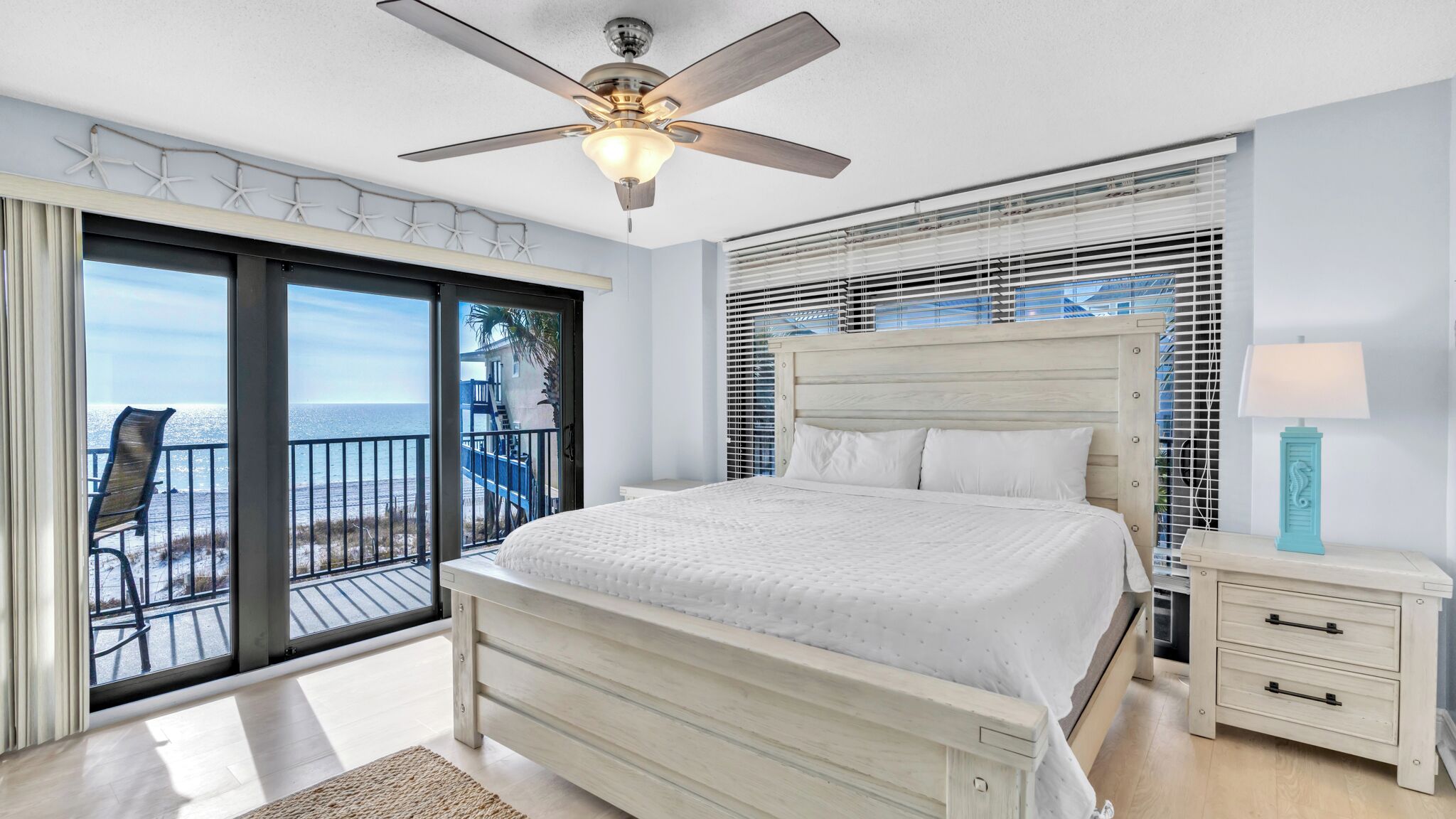 A bedroom with a bed, a nightstand, a ceiling fan, and a view of the ocean through large windows.