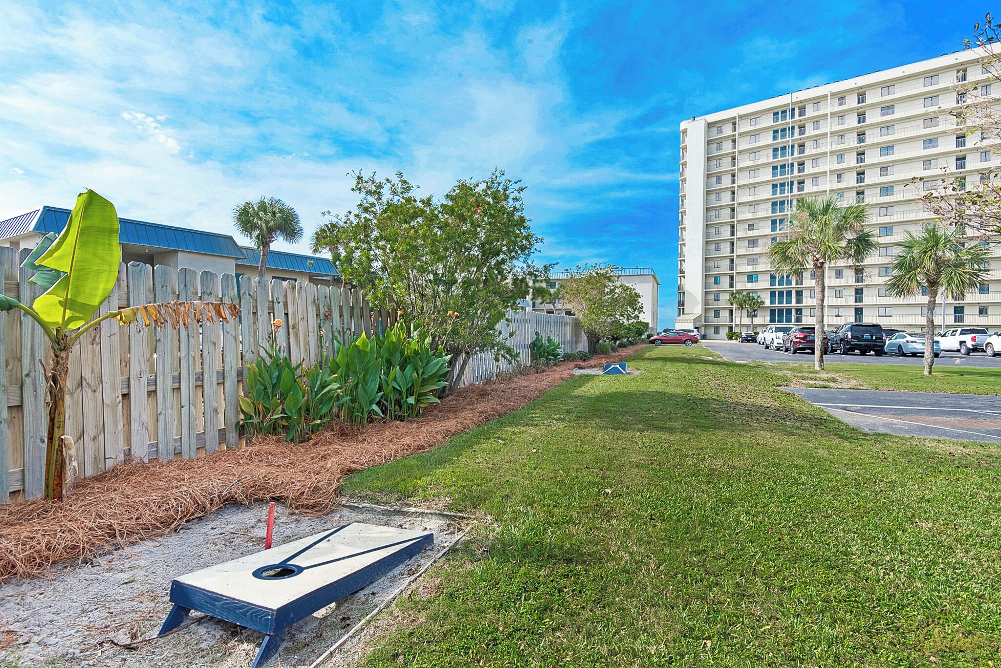 A grassy area with a shuffleboard table, a wooden fence, and a tall building in the background.