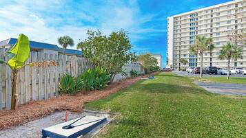 A grassy area with a shuffleboard table, a wooden fence, and a tall building in the background.