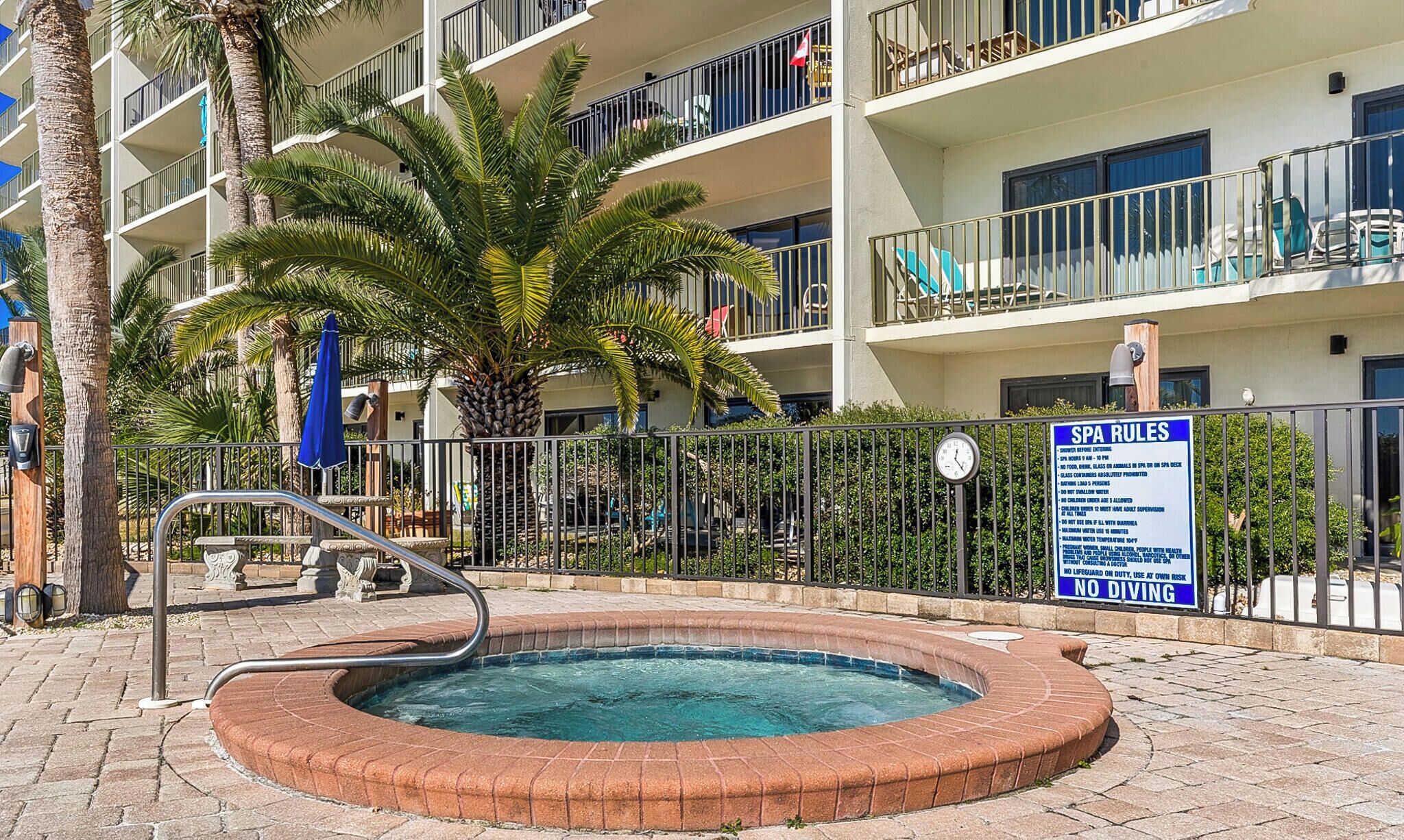 Una piscina con borde de ladrillo, rodeada por una valla metálica, palmeras y un edificio de varios pisos con balcones.