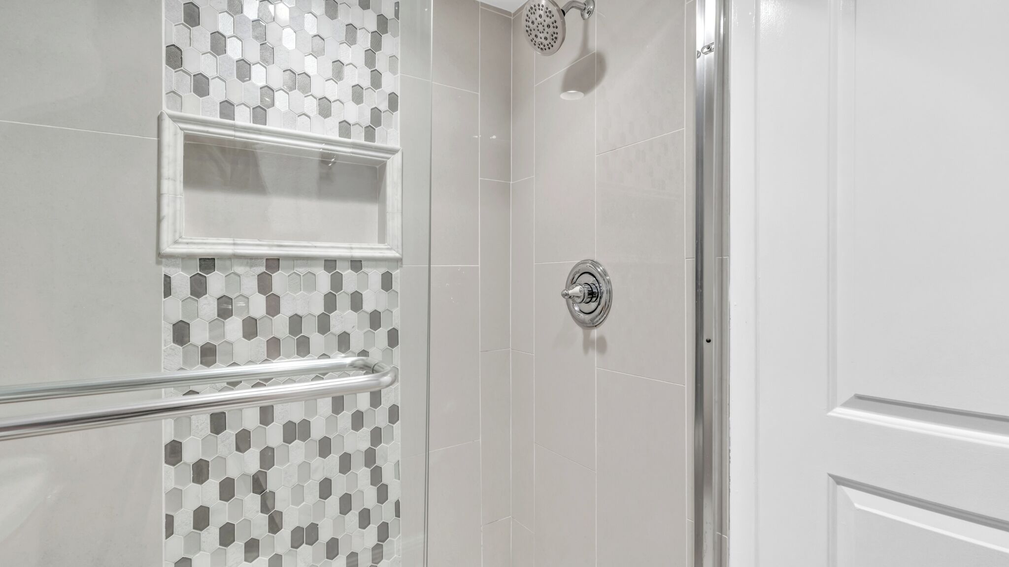 Une douche avec une porte en verre, un mur recouvert de carreaux blancs formant des motifs hexagonaux et une pomme de douche chromée.