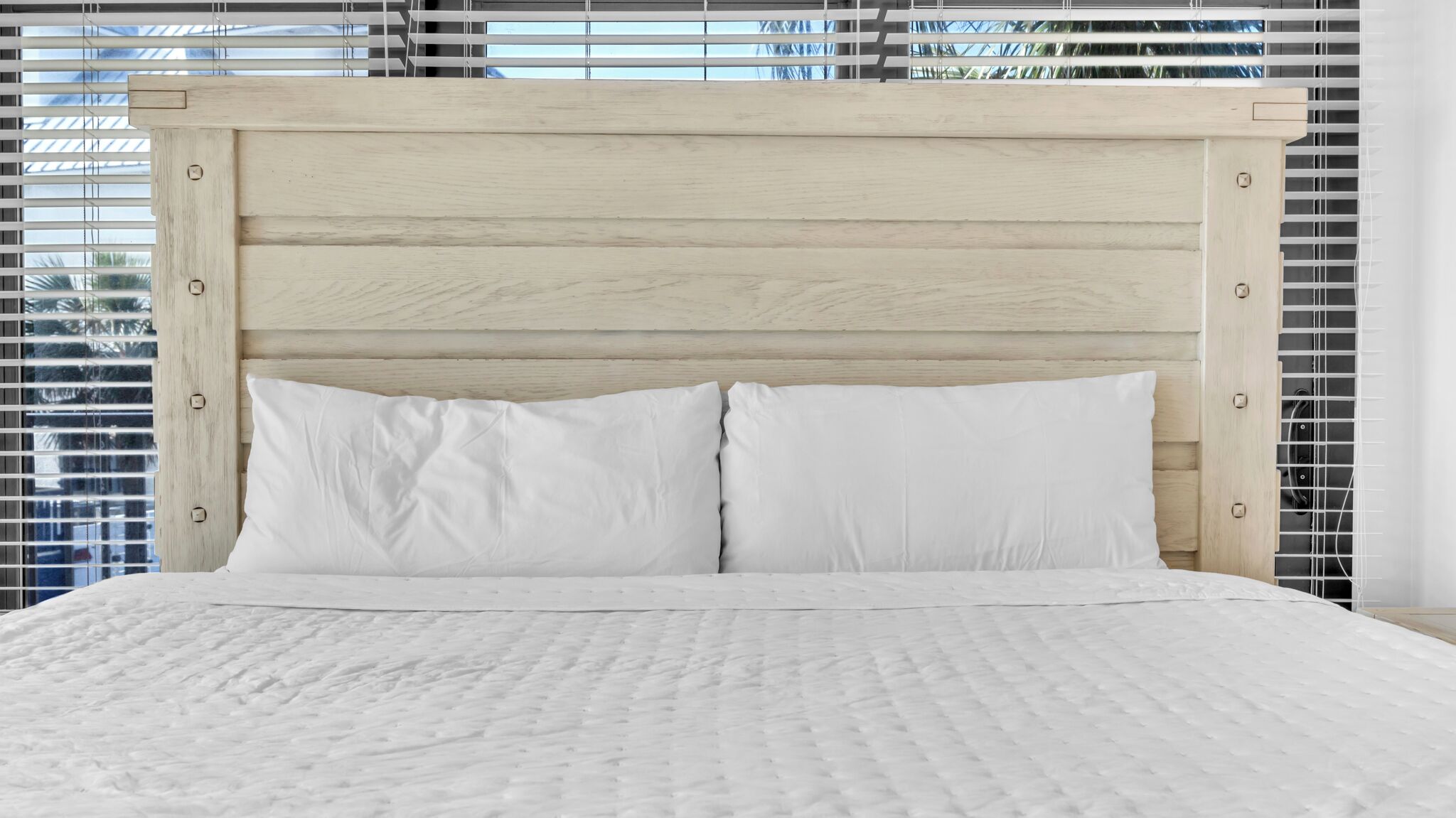 A bed with a wooden headboard, white bedding, and a view through window blinds.