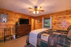 Dormitorio de una cabaña de madera con cama, cómoda, ventilador de techo y televisor.