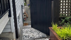 A narrow pathway leading to a gated area with a wooden fence, gravel steps, and a planter with green plants. - Grate Location in Upper Lansdale North Vancouver (North Vancouver)