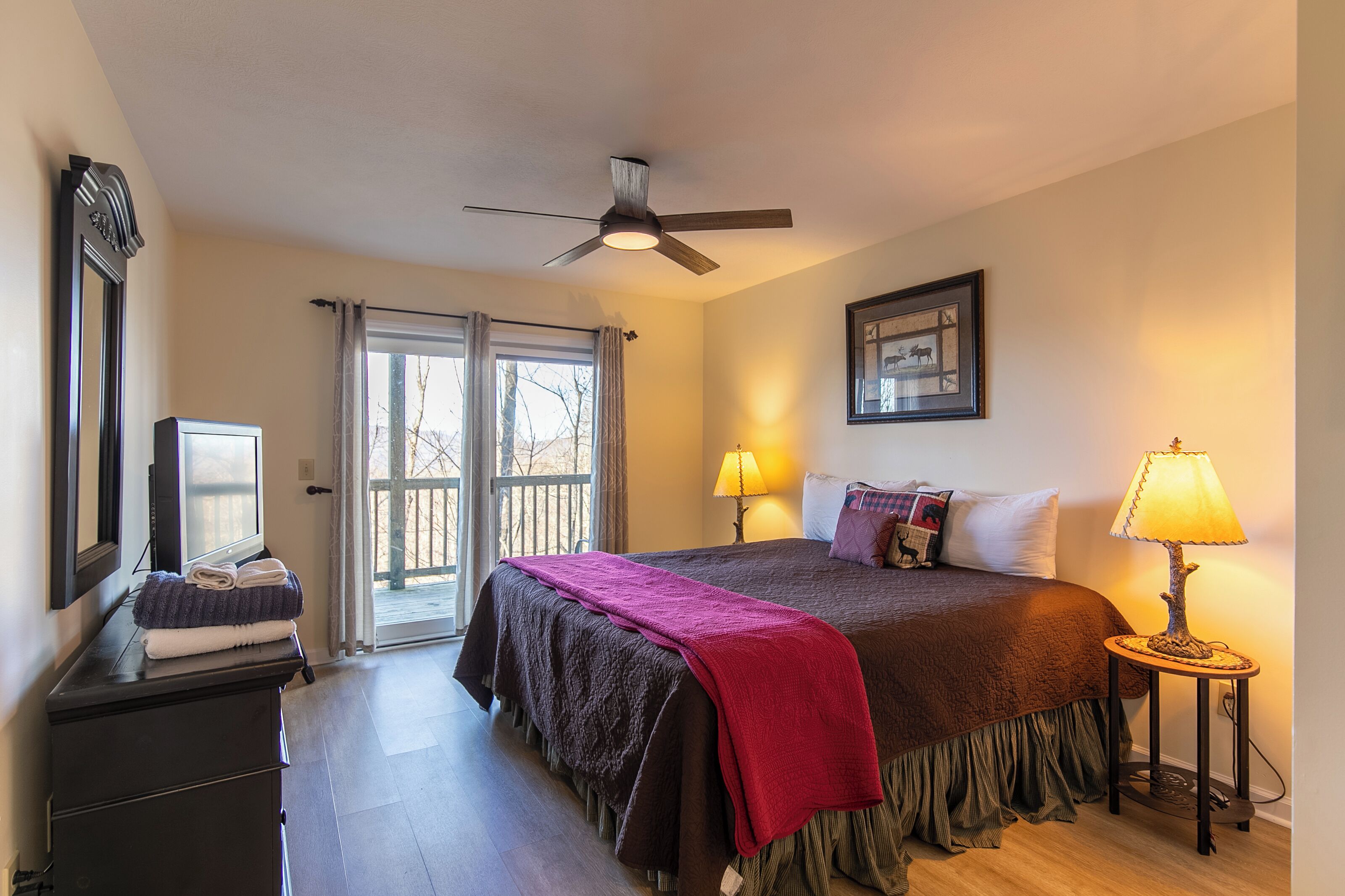 A hotel room with a bed, two bedside lamps, a ceiling fan, a dresser, a mirror, and a window with curtains.