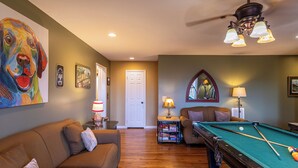 A living room with a pool table, a sofa, a ceiling fan, and a painting of a dog.