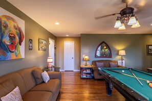 A living room with a pool table, a sofa, a ceiling fan, and a painting of a dog.